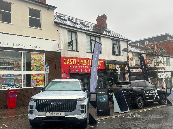 Wet urban street with parked cars near shops including a mini market and beauty store.