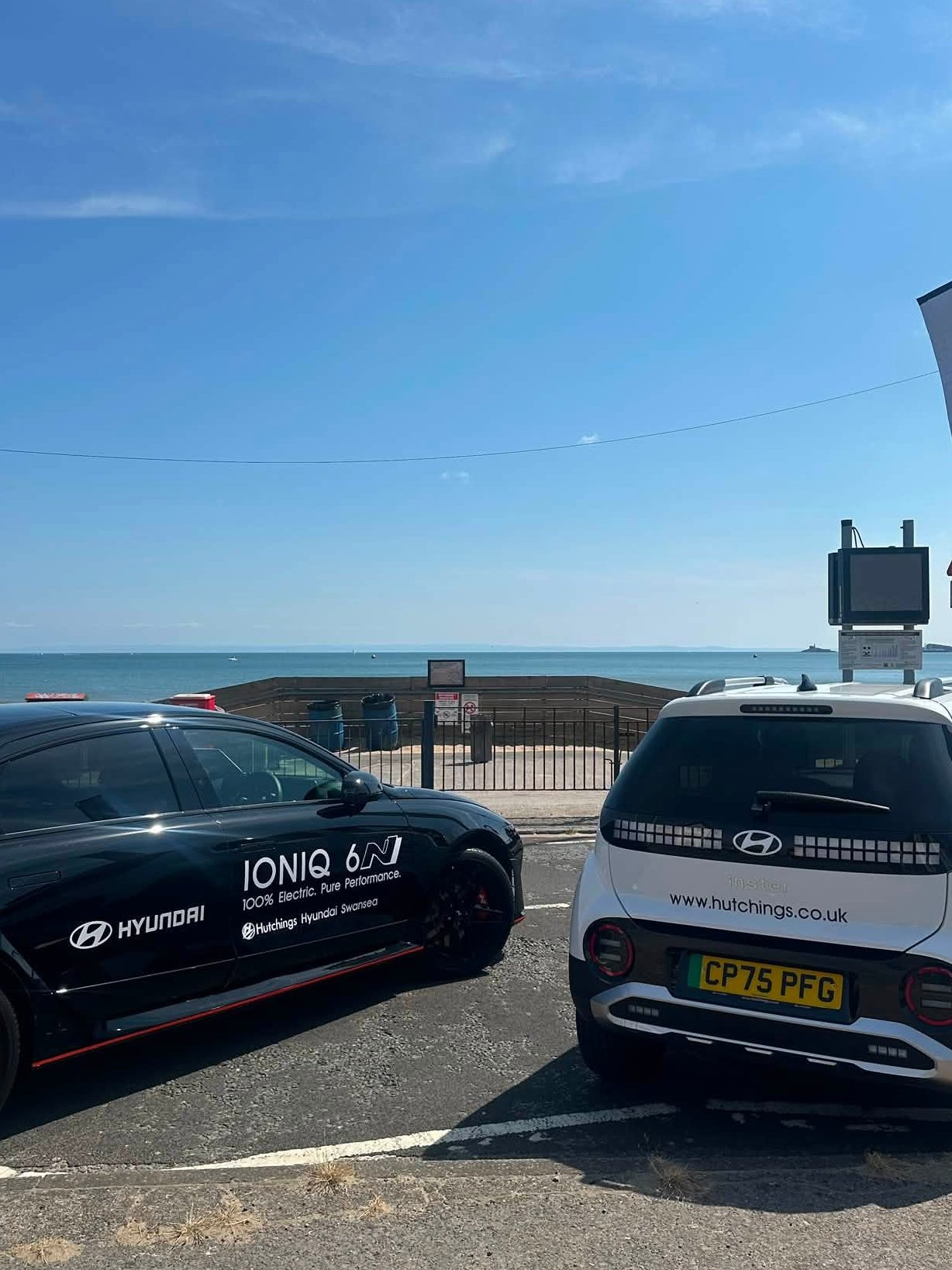 Two Hyundai cars parked near the sea, one black and one white, with a Hyundai flag nearby.