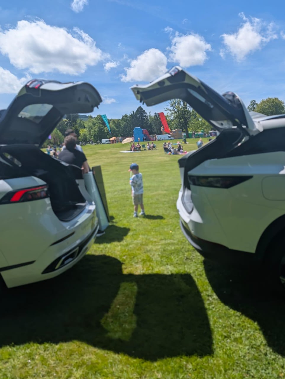 Two cars with open boots in a park setting, with people and colourful flags in the distance.