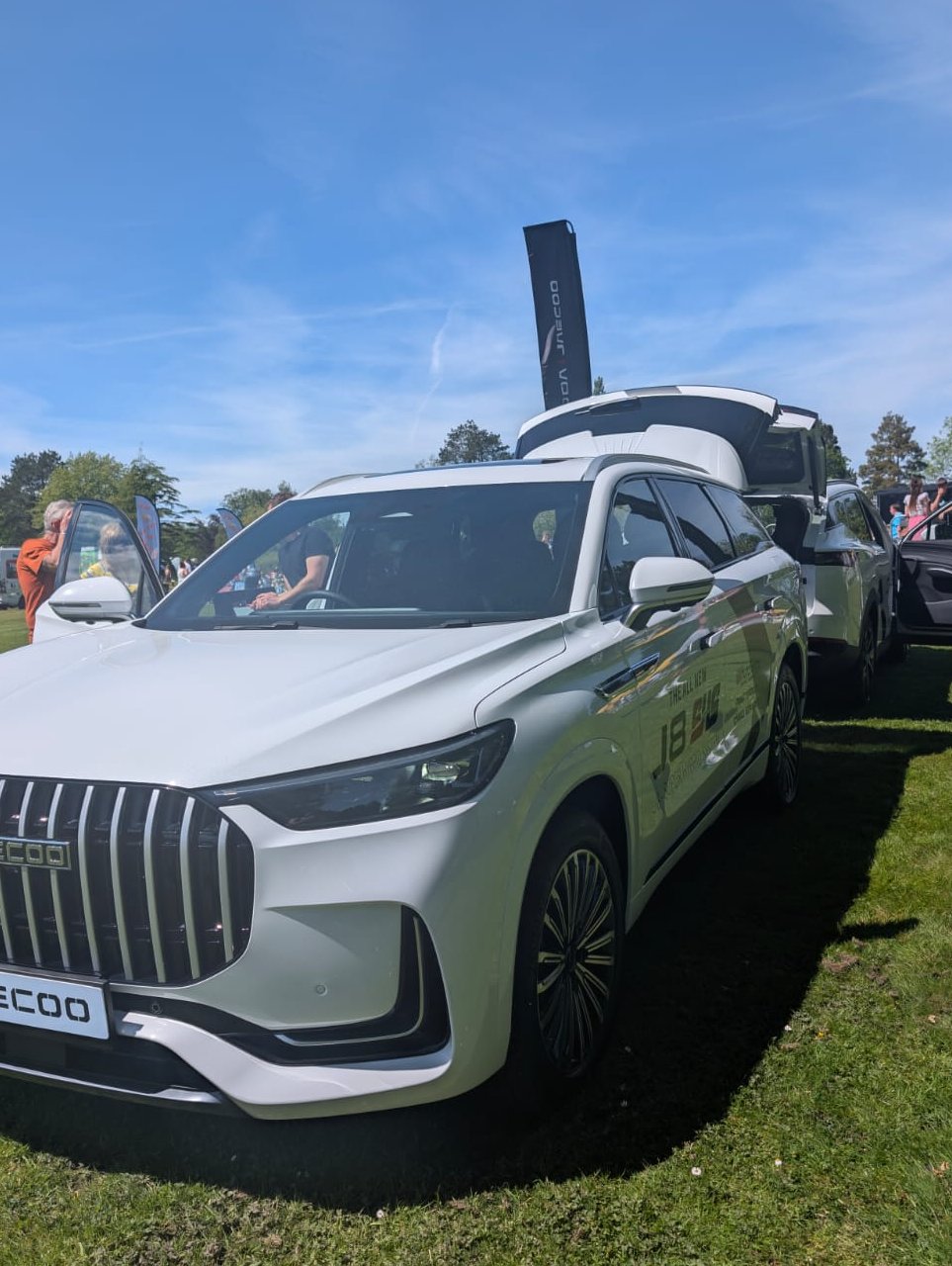 White Jaecco vehicle on a grass field with doors open and banners in background.