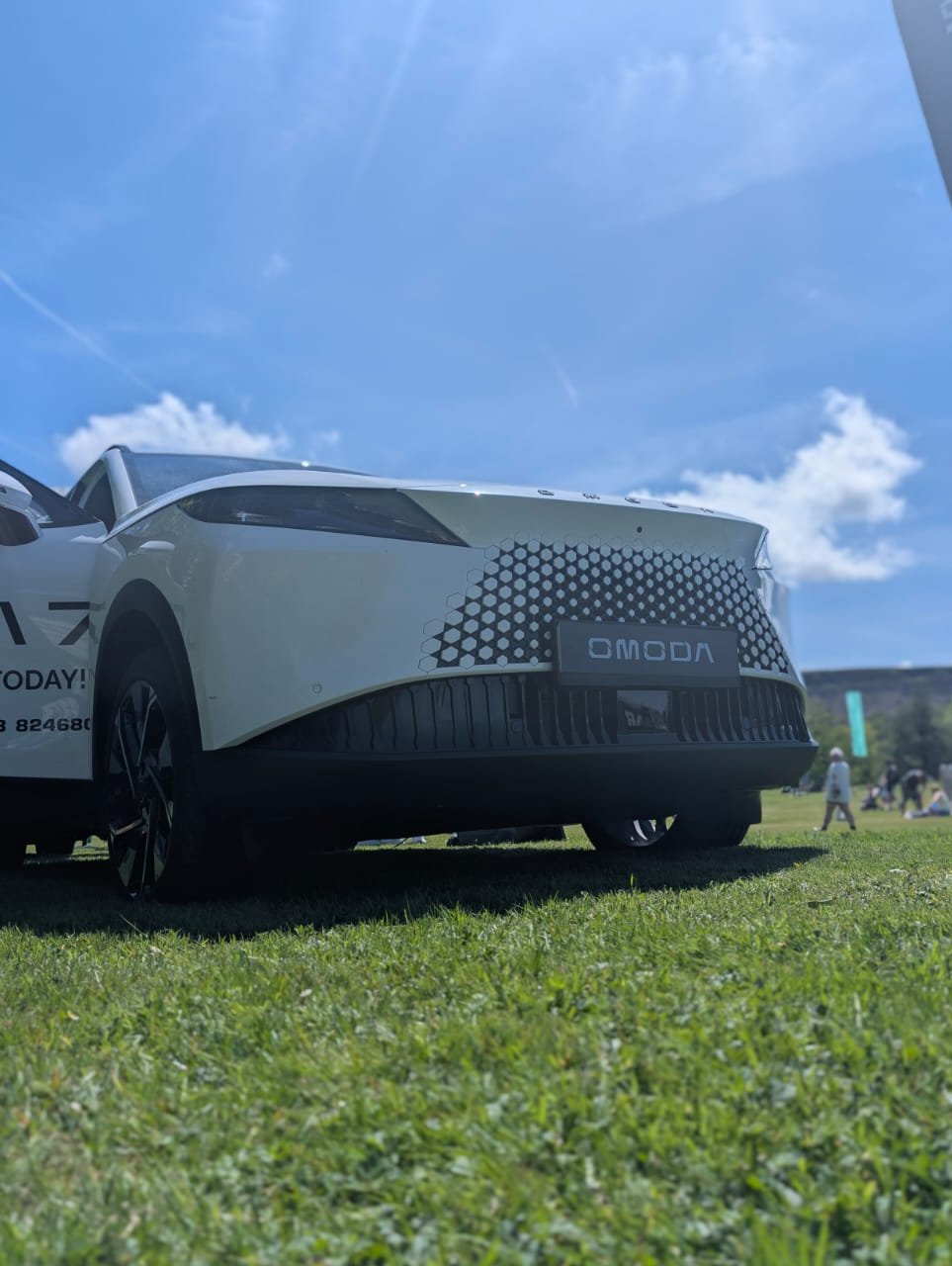 White OMODA car parked on grass with branded flag nearby.