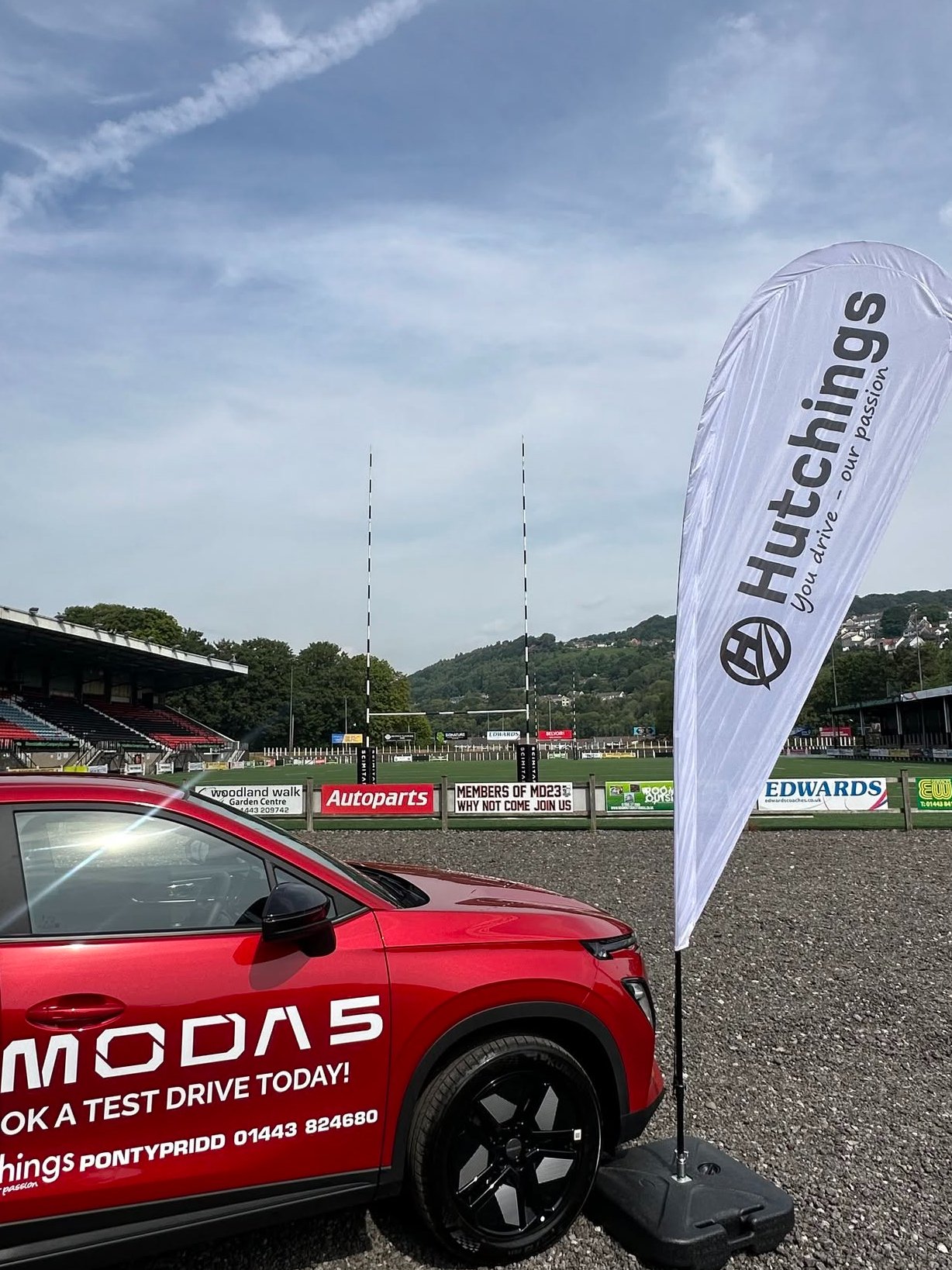Red Omoda 5 car with Hutchings flag next to a sports field.