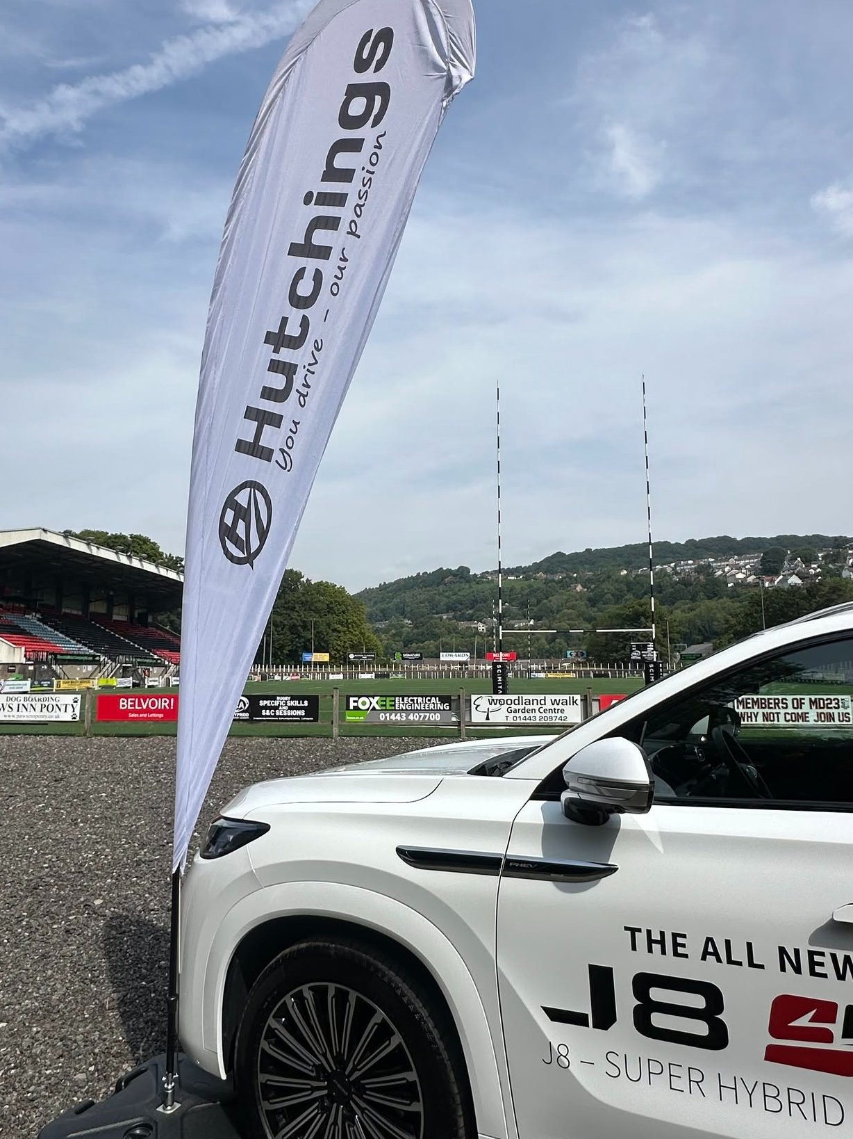 White car next to a Hutchings promotional flag at a sports ground.