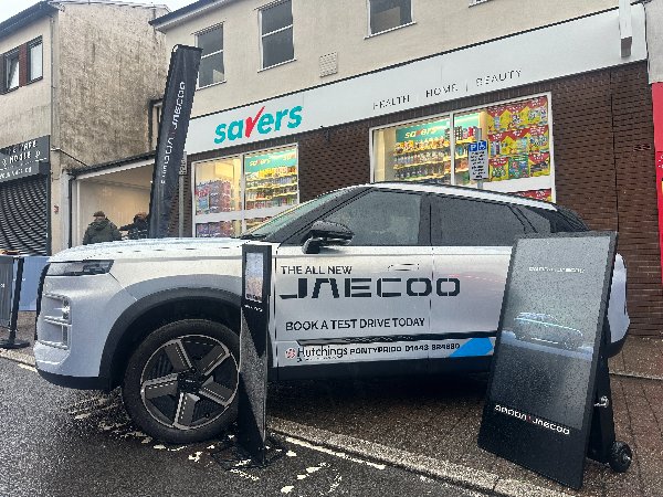 White vehicle with JAECOO branding parked outside a shop displaying a promotion board.