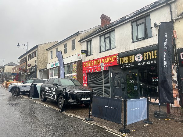 Two parked SUVs with OMODA branding next to shops on a rainy street.