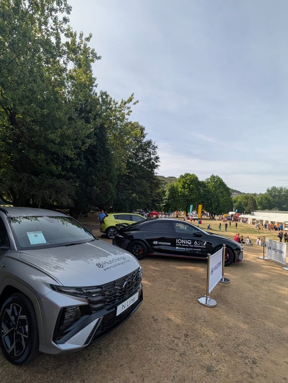 Silver car and black car displayed outdoors with trees in the background.
